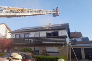 Ein Einfamilienhaus, dessen Dachstuhl durch einen Brand schwer beschädigt wurde. Im Vordergrund Feuerwehrleute und eine Drehleiter der Feuerwehr.